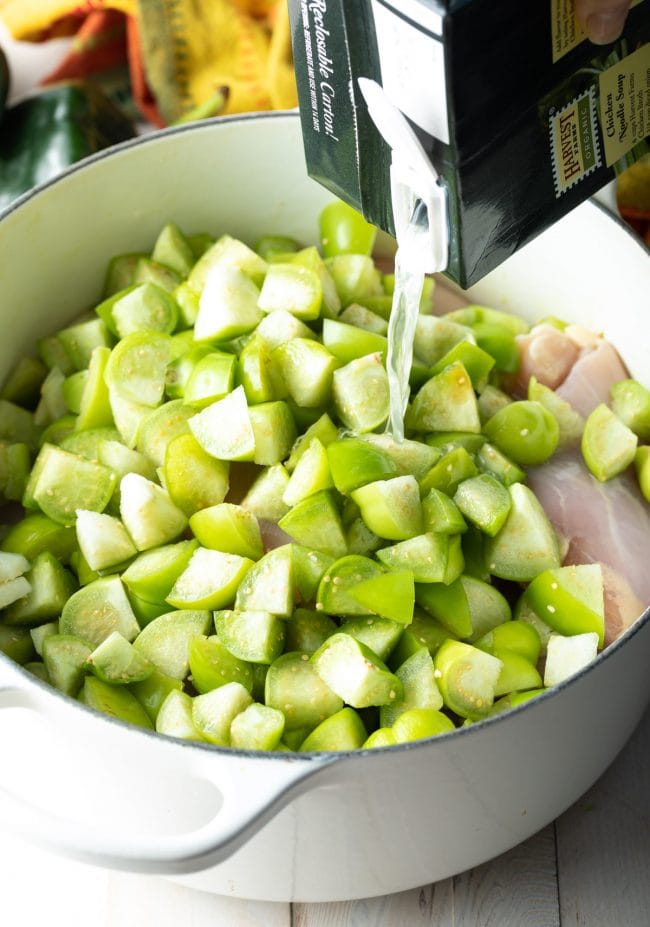 Tomatillos and chicken in a pot with a hand pouring chicken broth over the top. 