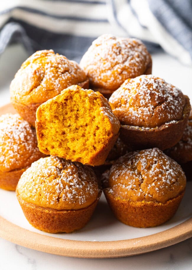 Stack of fluffy muffins made with pumpkin puree, each dusted with powdered sugar. Top muffin has been bitten in half to show inside.