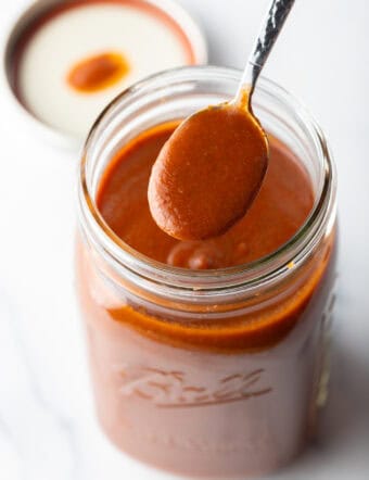 Spoonful of red enchilada sauce being held to camera over a large jar.