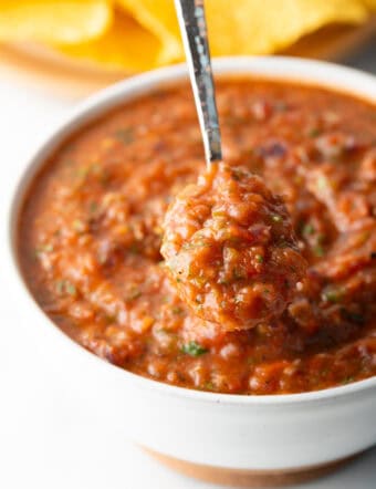 A metal spoon is showing a scoop of roasted salsa scooped from a white bowl.