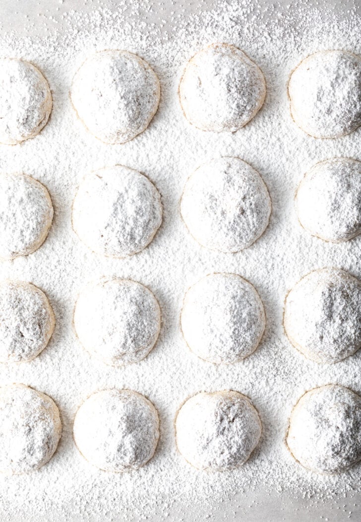 Top down view 16 snowball cookies on a baking sheet, covered with a heavy layer of powdered sugar.