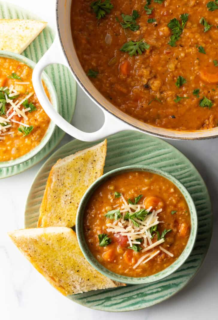 Top down view green bowl of red lentil soup topped with chopped herbs and shredded cheese. The bowl is on a plate with two baguette slices.