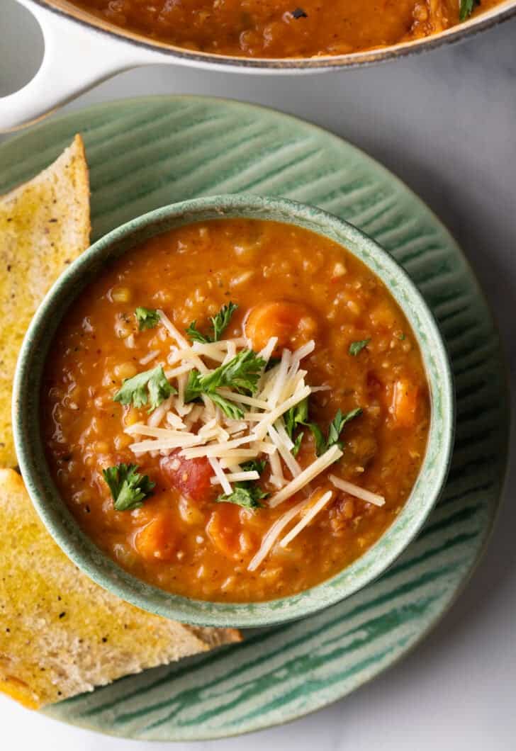 Top down view green bowl of red lentil soup topped with chopped herbs and shredded cheese. The bowl is on a plate with two baguette slices.