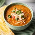 Green bowl of red lentil soup topped with chopped herbs and shredded cheese. The bowl is on a plate with two baguette slices.