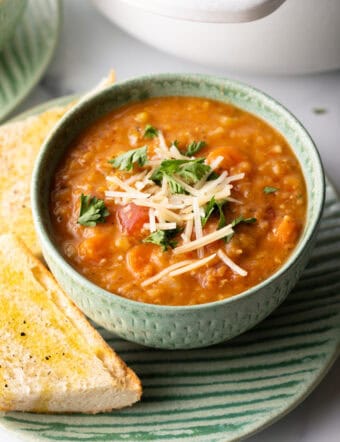 Green bowl of red lentil soup topped with chopped herbs and shredded cheese. The bowl is on a plate with two baguette slices.