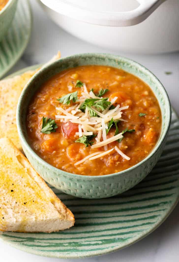 Green bowl of red lentil soup topped with chopped herbs and shredded cheese. The bowl is on a plate with two baguette slices.