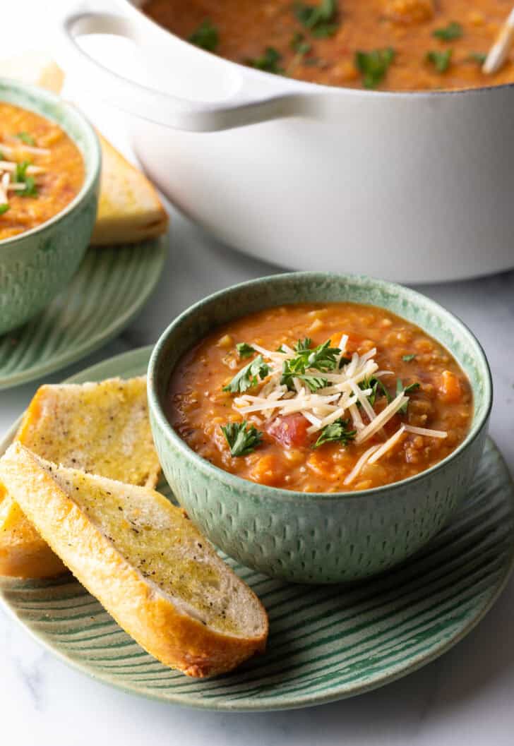 Green bowl of red lentil soup topped with chopped herbs and shredded cheese. The bowl is on a plate with two baguette slices.