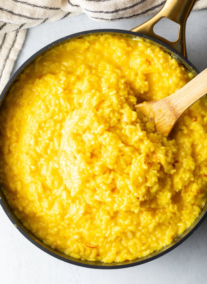 Wooden spatula stirring yellow risotto in a large deep skillet.