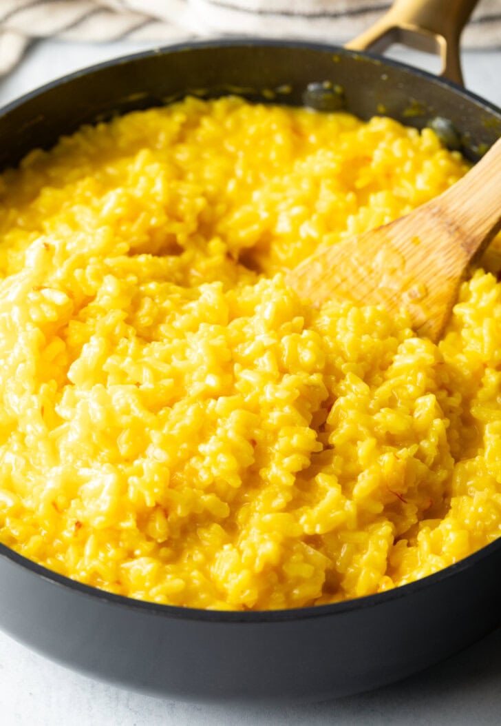 Wooden spatula stirring yellow risotto in a large deep skillet.