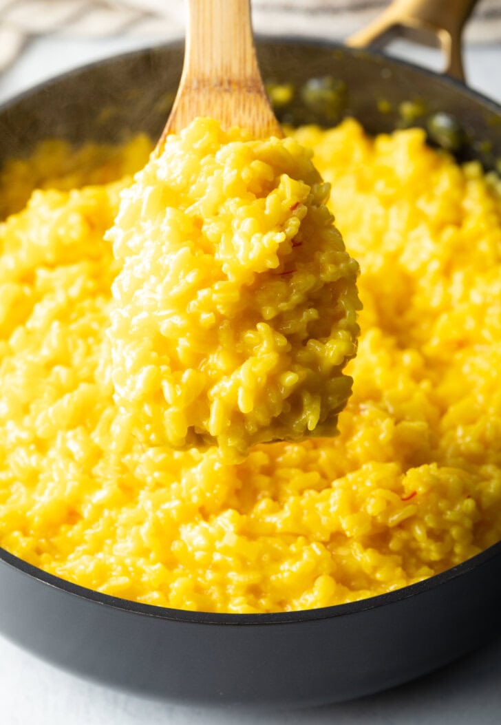 Wooden spatula holding a serving of yellow risotto above a large deep skillet, showing to camera.