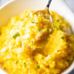 Metal spoon scooping a bite of yellow risotto milanese from a white bowl, showing to camera.