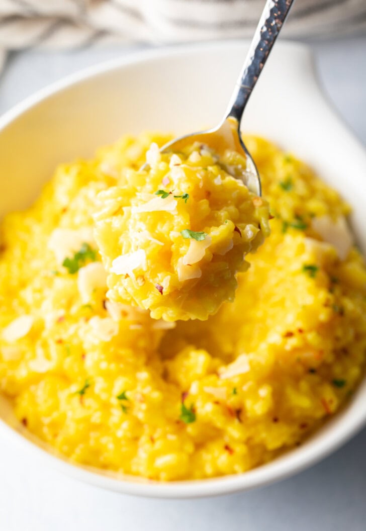 Metal spoon scooping a bite of yellow risotto milanese from a white bowl, showing to camera.