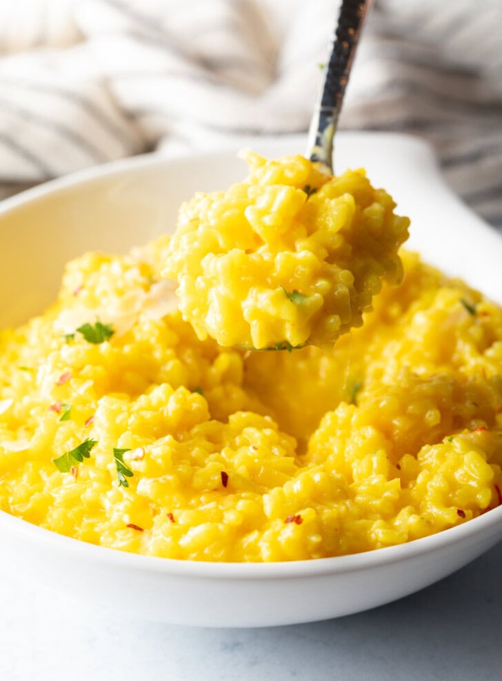 Metal spoon scooping a bite of yellow risotto milanese from a white bowl, showing to camera.