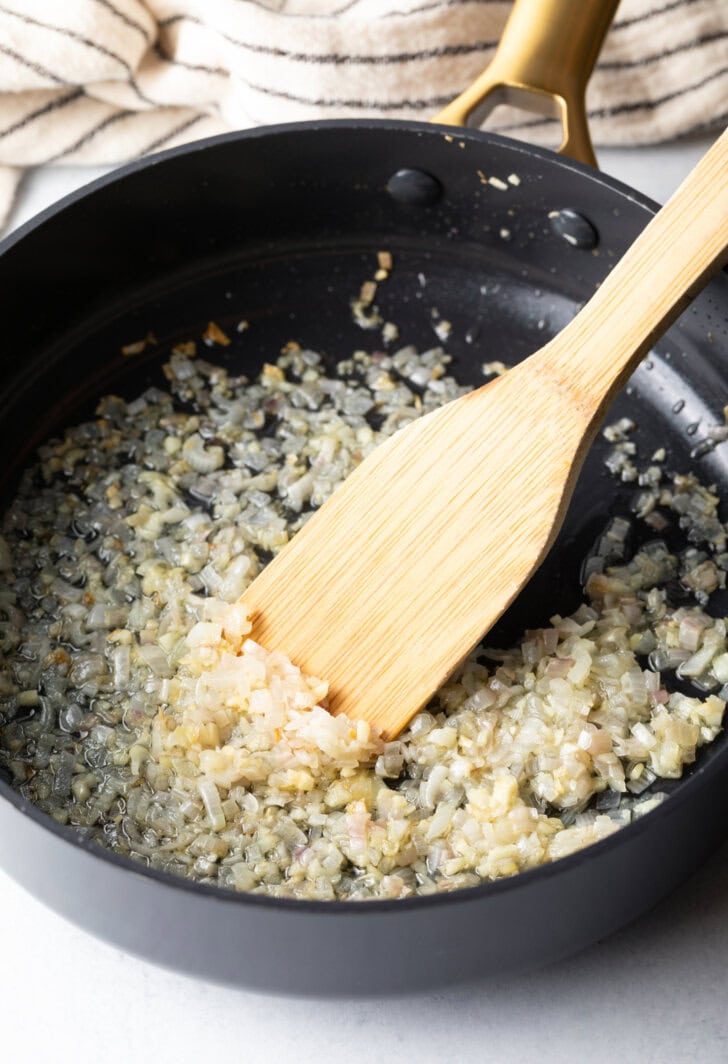 Wooden spatula sauteeing onions and garlic in a large deep skillet.