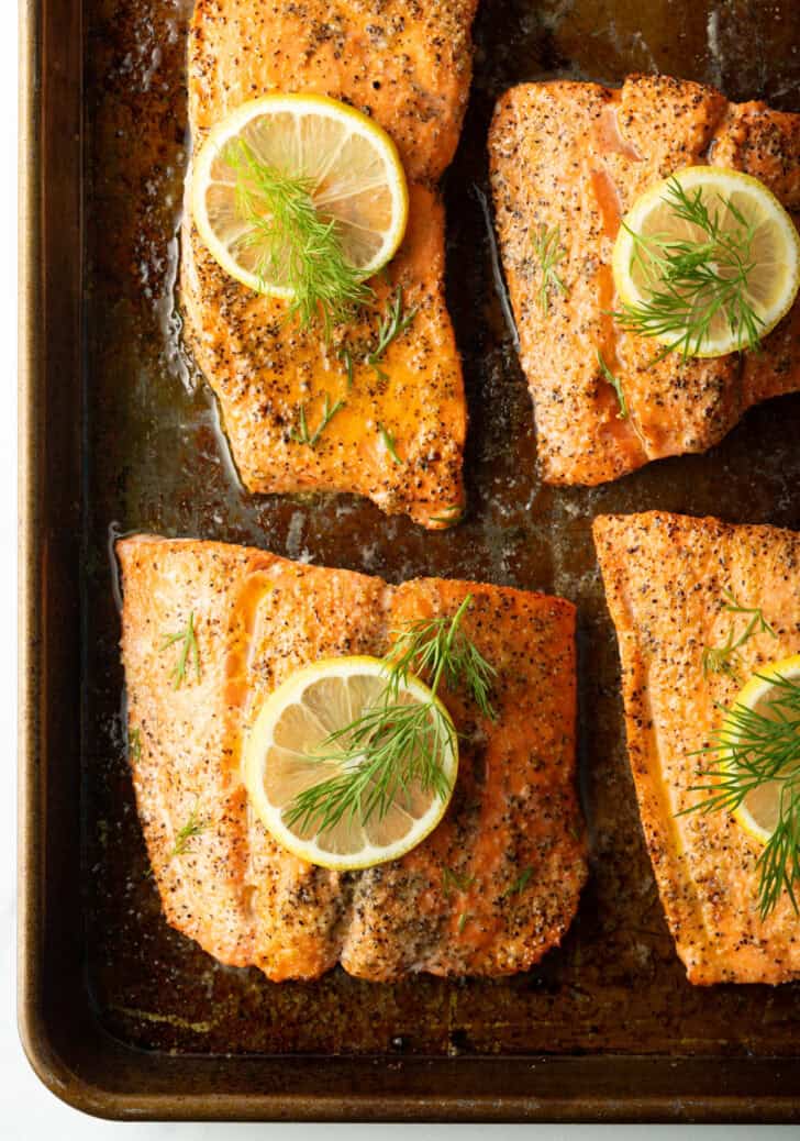 Top down view 4 large baked salmon filets on a baking sheet. Each piece is topped with a slice of fresh lemon and dill sprigs.