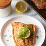 Top down view of a large filet of salmon topped with a lemon slice and sprigs of fresh dill. The fish is on a white plate.