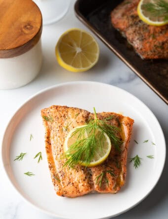 Top down view of a large filet of salmon topped with a lemon slice and sprigs of fresh dill. The fish is on a white plate.