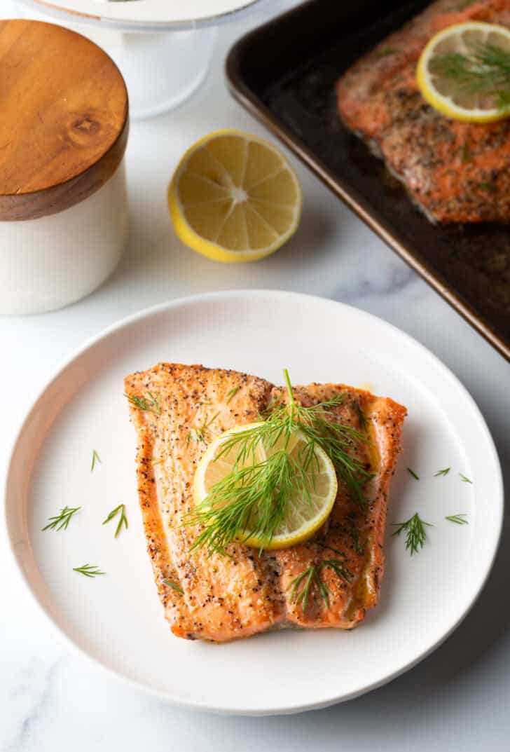 Top down view of a large filet of salmon topped with a lemon slice and sprigs of fresh dill. The fish is on a white plate.