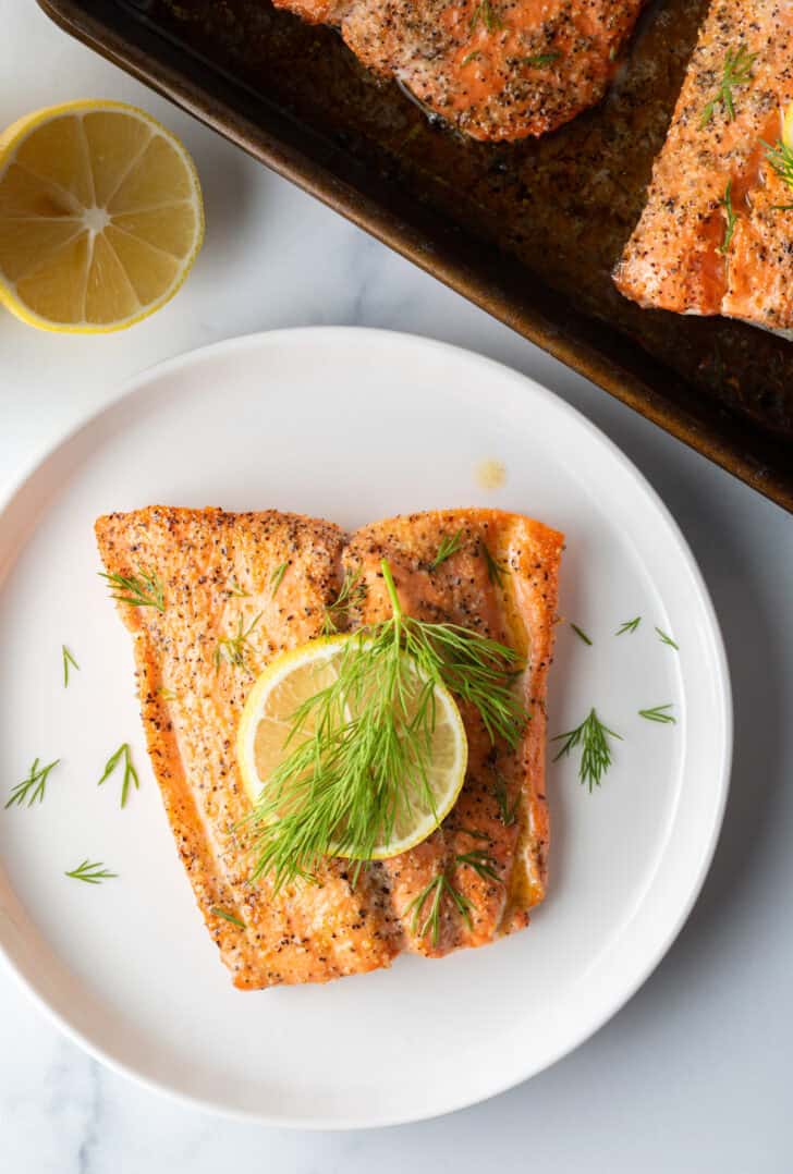 Top down view of a large filet of salmon topped with a lemon slice and sprigs of fresh dill. The fish is on a white plate.