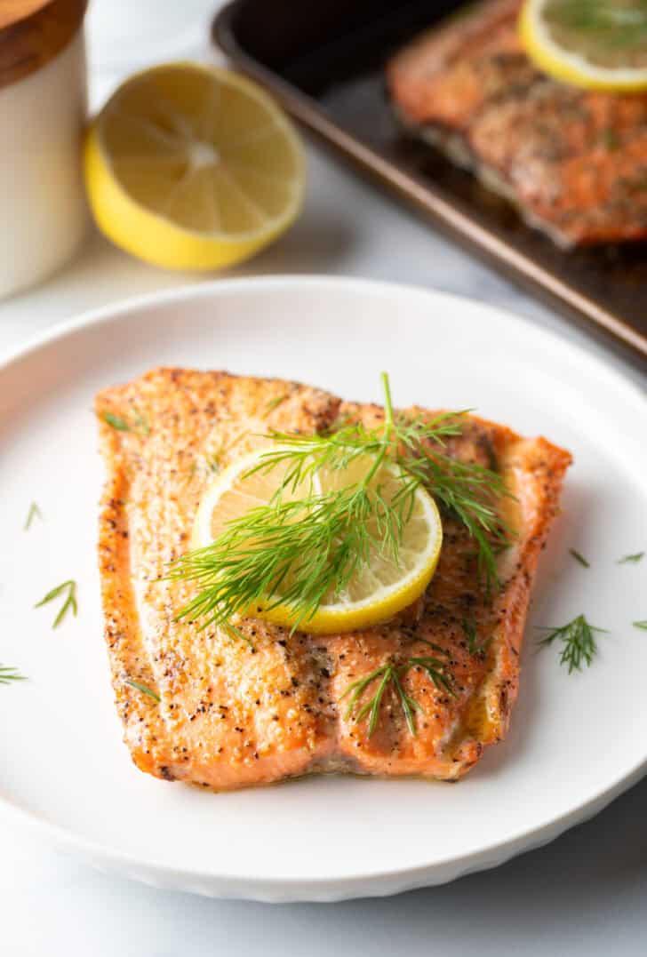 Large filet of salmon topped with a lemon slice and sprigs of fresh dill. The fish is on a white plate.