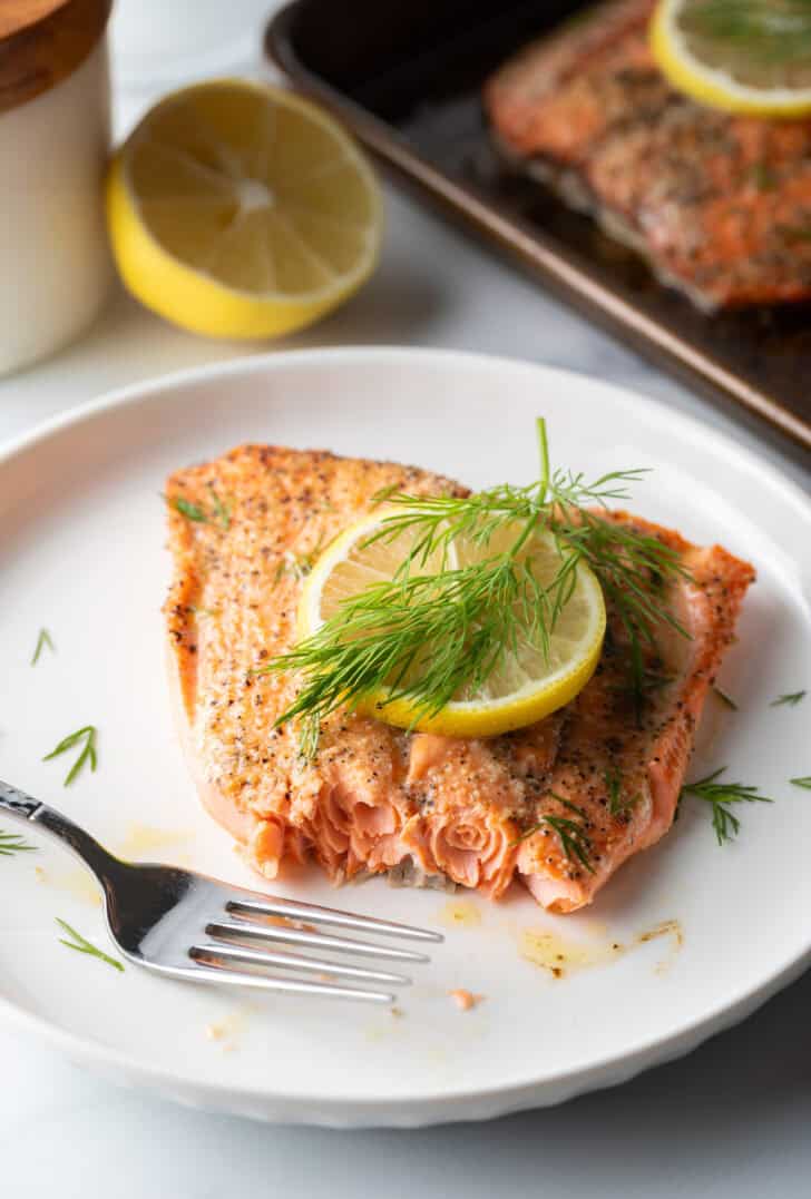 Large filet of salmon topped with a lemon slice and sprigs of fresh dill. The fish is on a white plate. A fork has removed a bite from the salmon.