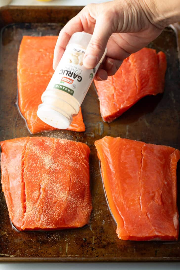 Hand with a salt shaker adding spices onto 4 pieces of raw salmon on a baking sheet.