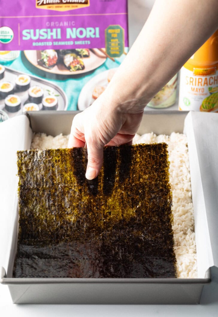 Hand adding large sheet of nori on top of the rice in a rectangle baking pan.