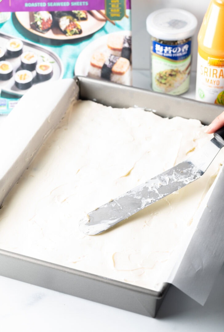 Using a flat metal spatula to smooth cream cheese into a rectangle baking pan.