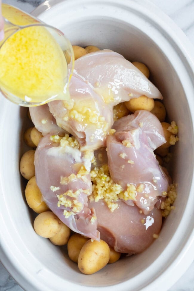 Raw chicken and potatoes in a slow cooker with a garlic butter mixture being poured over the top. 