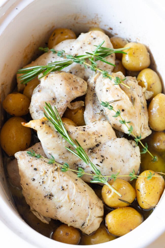 Slow cooker with small potatoes and chicken in it topped with fresh herbs. 