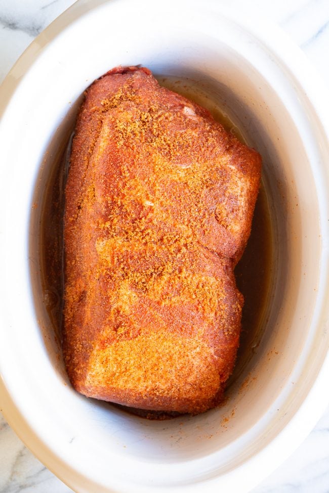 Pork butt roast in a white crockpot with brown sugar and seasoning rubbed on top.