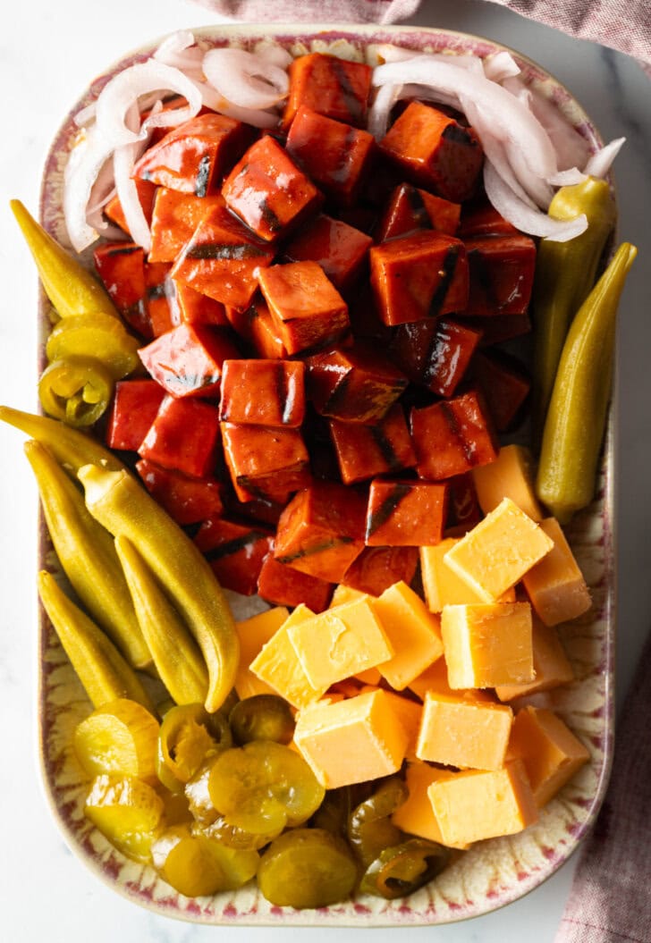 Top down view of a serving plate with a stack of bbq smoked bologna cubes, squares of cheese, pickled okra, slices of red onion, and pickles.