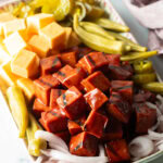 Serving plate with a stack of bbq smoked bologna cubes, squares of cheese, pickled okra, slices of red onion, and pickles.