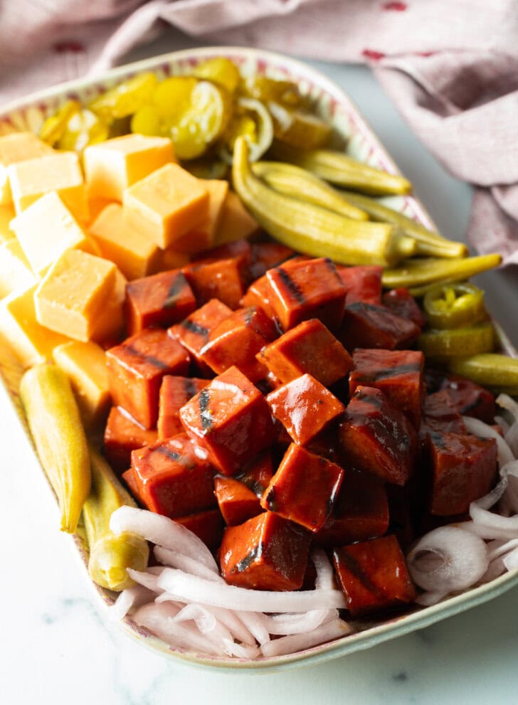 Serving plate with a stack of bbq smoked bologna cubes, squares of cheese, pickled okra, slices of red onion, and pickles.