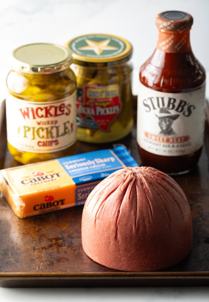 Recipe ingredients on a baking tray: Jar of wickles pickles, jar of pickled okra, a block of cheddar cheese, the butt end of a cut of bologna, and a bottle of bbq sauce.