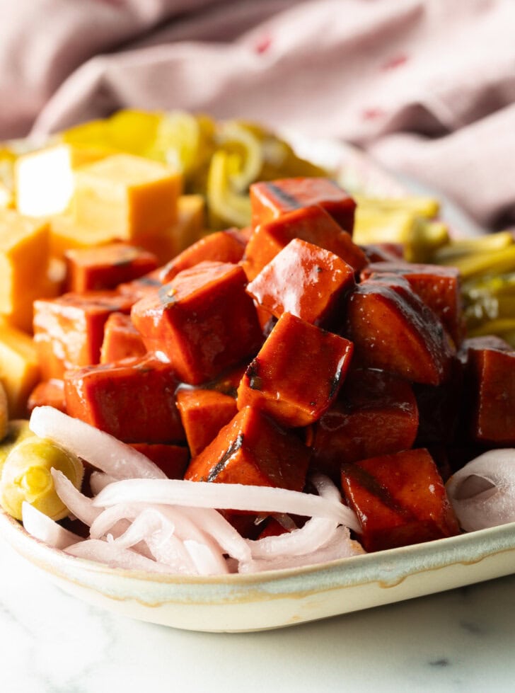 Serving plate with a stack of bbq smoked bologna cubes, squares of cheese, pickled okra, slices of red onion, and pickles.