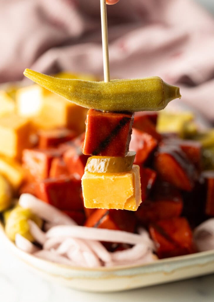 Toothpick stacked with a cube of cheddar cheese, cube of smoked bologna, and an okra spear, held to camera.