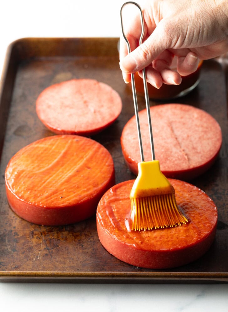 Hand using a yellow basting brush to coat 4 thick slice of baloney with barbecue sauce.