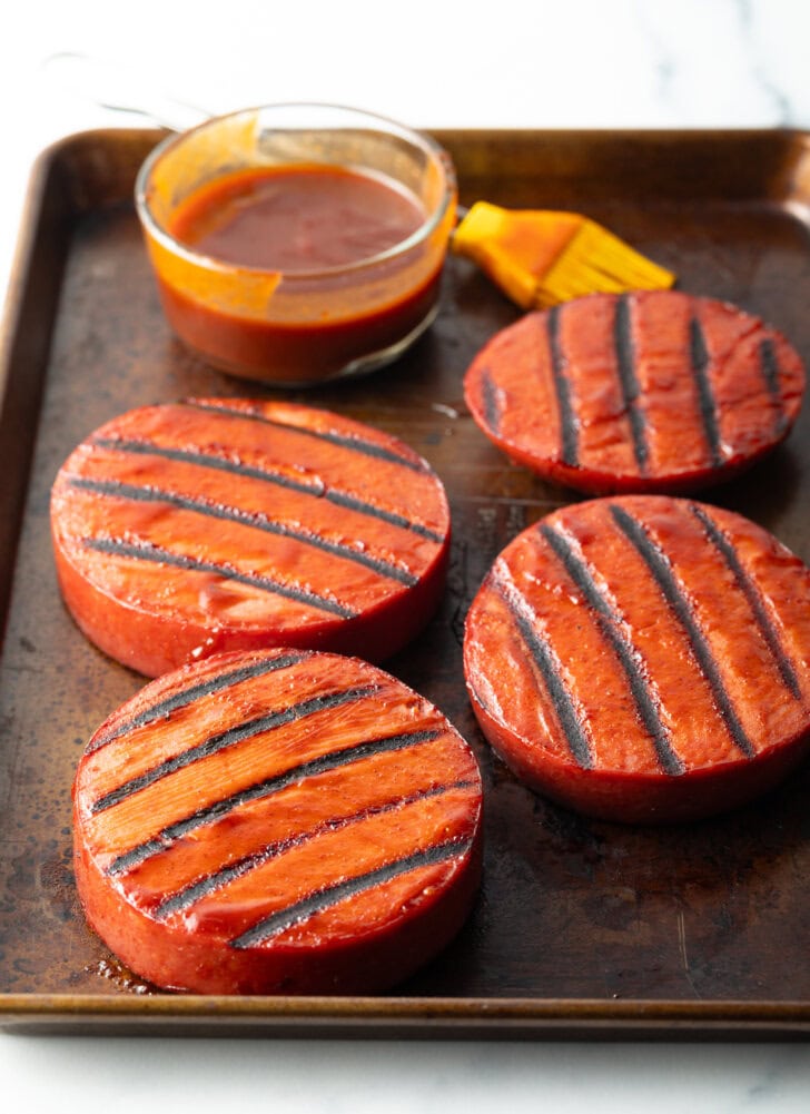 4 rounds of bologna with grill marks. In the background in a glass dish with bbq sauce and a yellow basting brush.