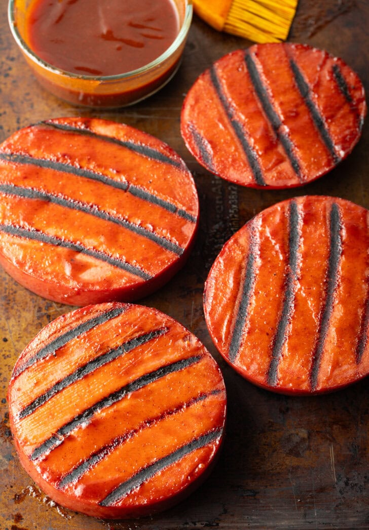 4 rounds of bologna with grill marks. In the background in a glass dish with bbq sauce and a yellow basting brush.