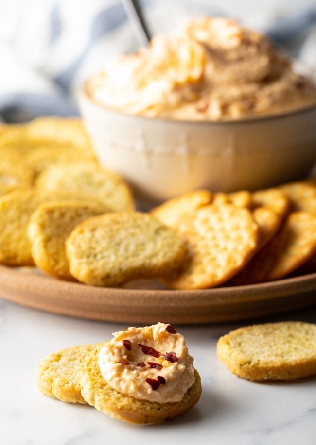 pieces of sliced bread, one topped with smoked cheese dip and platter with crackers and bowl of spread in the background