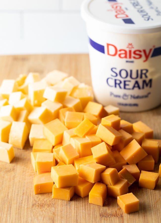 cubes of gouda and cheddar on a wood cutting board with container of sour cream in the background