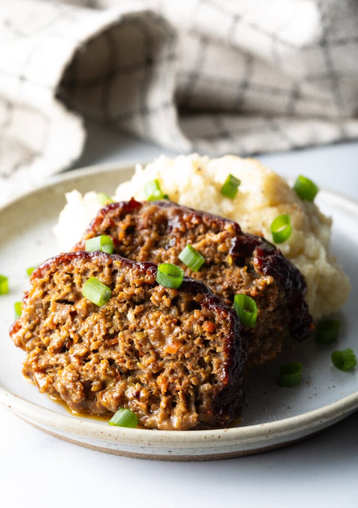 2 slices of meatloaf on a plate with a scoop of mashed potatoes, all topped with chopped green onions for garnish.