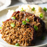 2 slices of meatloaf on a plate with a scoop of mashed potatoes, all topped with chopped green onions for garnish.