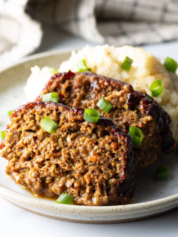 2 slices of meatloaf on a plate with a scoop of mashed potatoes, all topped with chopped green onions for garnish.