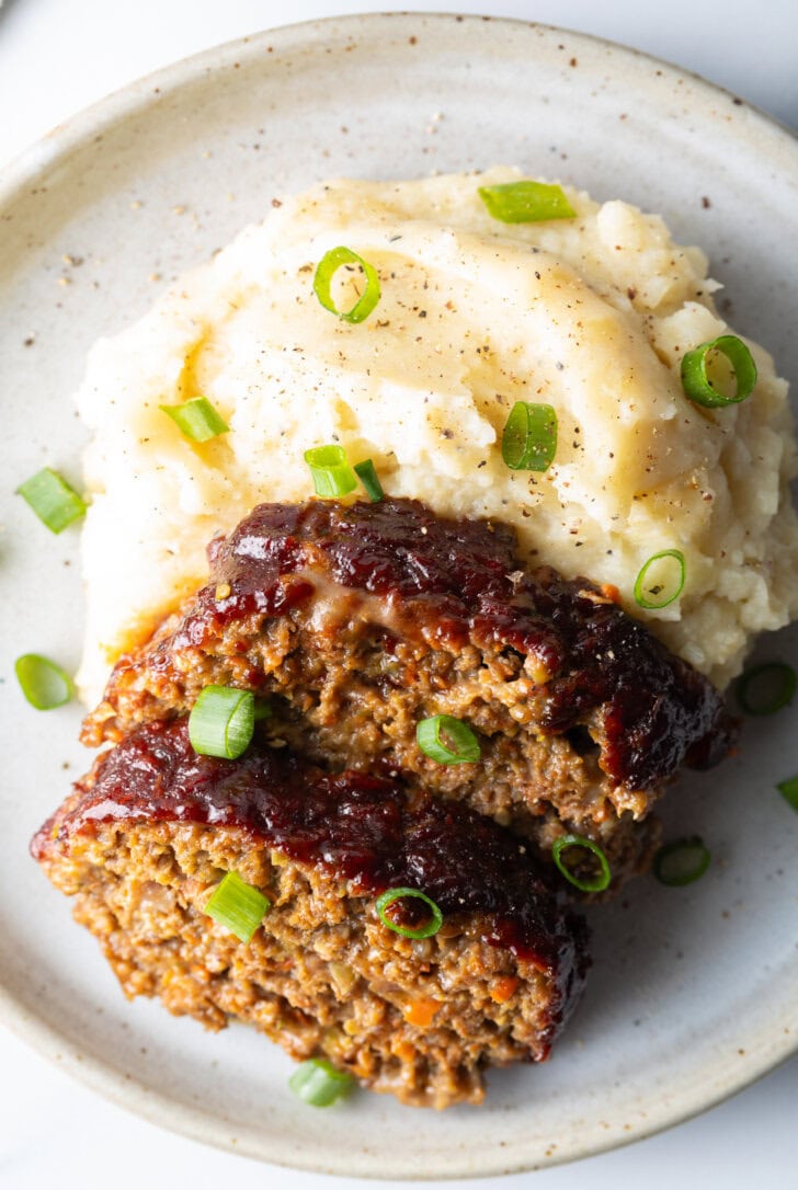 2 slices of meatloaf on a plate with a scoop of mashed potatoes, all topped with chopped green onions for garnish.