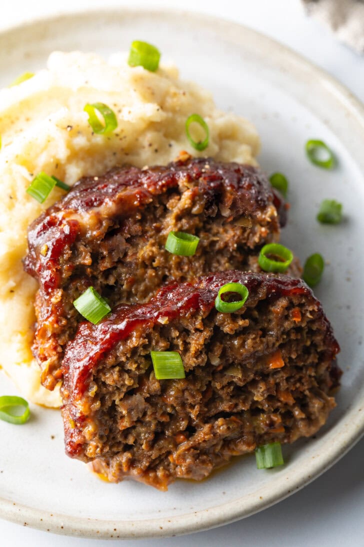 2 slices of meatloaf on a plate with a scoop of mashed potatoes, all topped with chopped green onions for garnish.