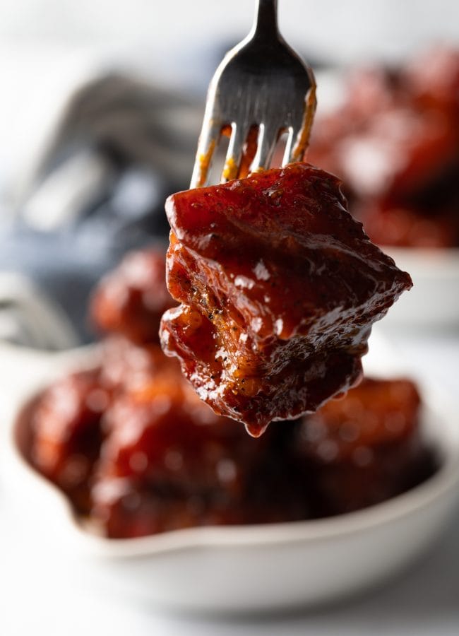 Fork holding a cube of smoked pork belly burnt ends recipes coated in bbq sauce to camera.