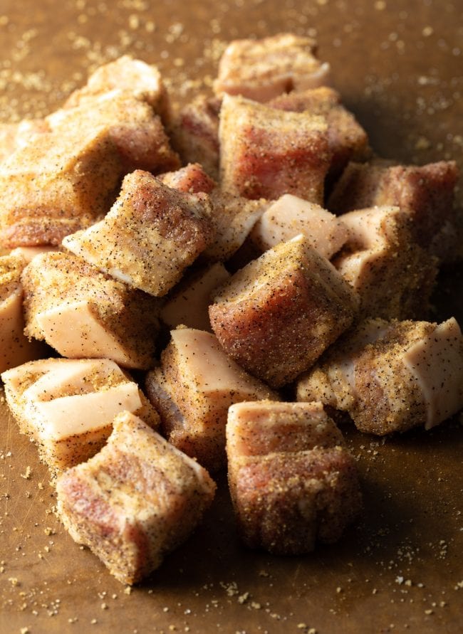 Cubes of raw pork tossed with spice rub on a cutting board.