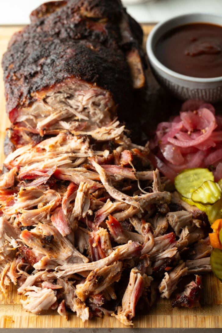 Shredded smoked pork butt on a cutting board, with ramekin of bbq sauce, stack of pickles, and pickled red onions.